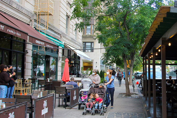 Two parents with three baby carriages are walking in front of an open restaurant